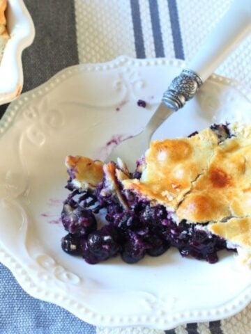 Slice of easy homemade blueberry pie on a white plate with golden crust and fresh, juicy blueberry filling, made with a store-bought crust.