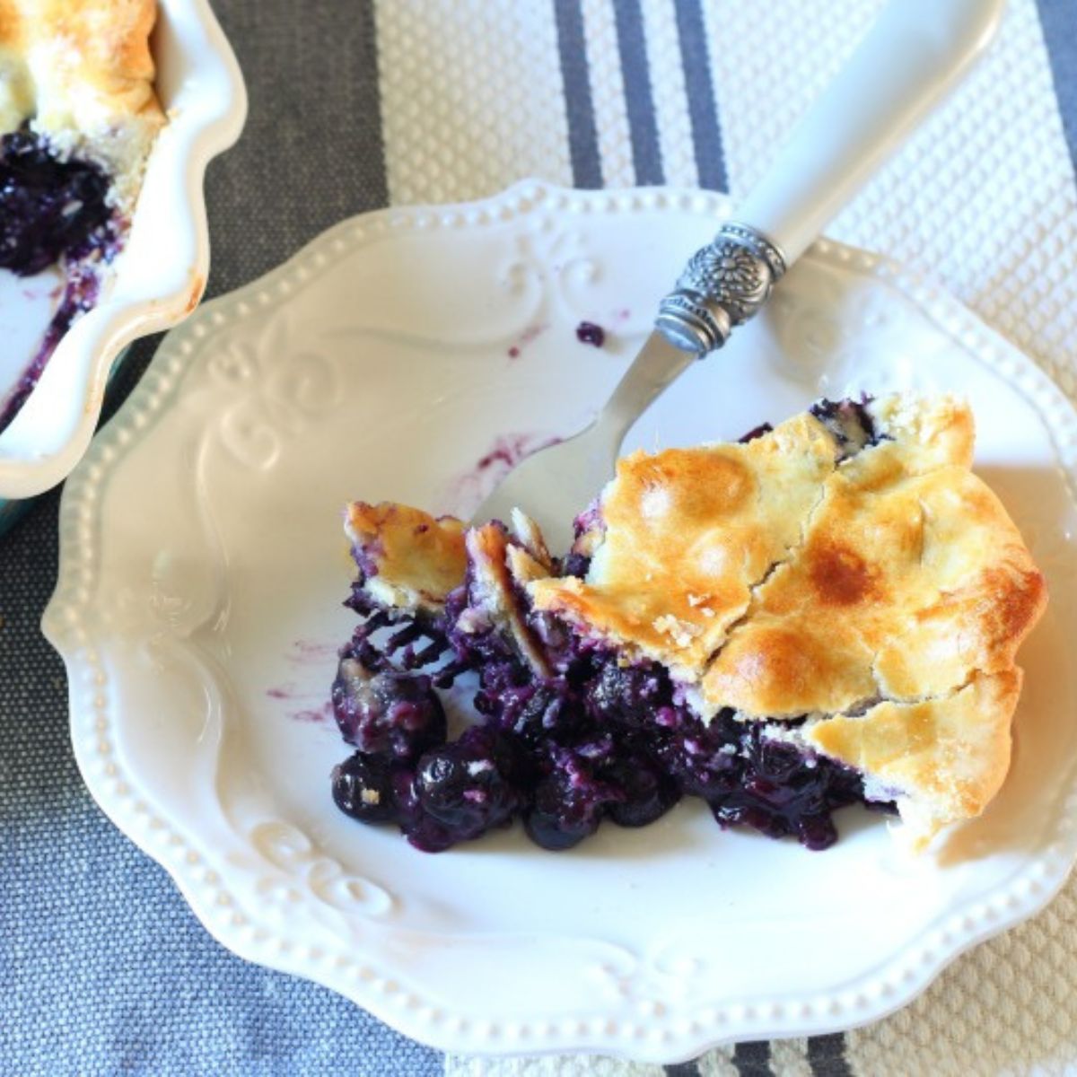 Slice of easy homemade blueberry pie on a white plate with golden crust and fresh, juicy blueberry filling, made with a store-bought crust.