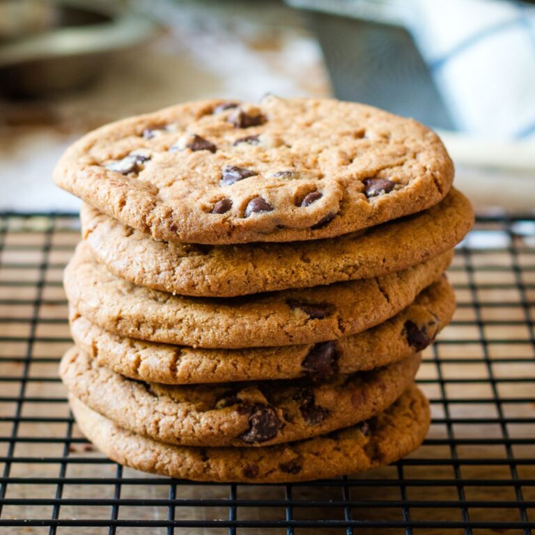 A stack of six large chocolate chip cookies, freshly baked and golden brown, arranged neatly on a cooling rack with a blurred kitchen background.