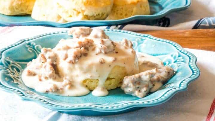 Side view of a blue plate with a fluffy biscuit on it topped with homemade sausage gravy with plate of fresh biscuits in the background
