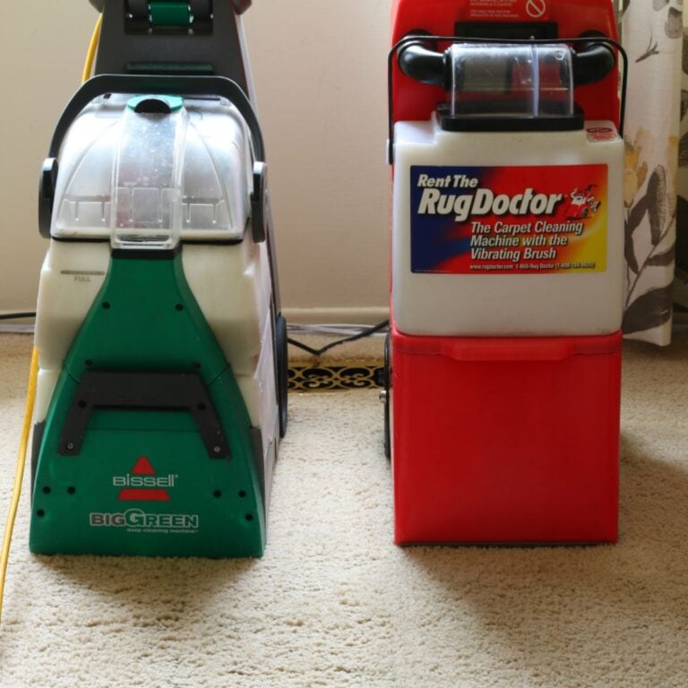 A side-by-side comparison of two carpet cleaning machines on a beige carpet. On the left is the Bissell Big Green Deep Cleaning Machine, which is primarily green with a clear water tank. On the right is the Rug Doctor carpet cleaning machine, which is red with a white and blue label that says "Rent The Rug Doctor - The Carpet Cleaning Machine with the Vibrating Brush.