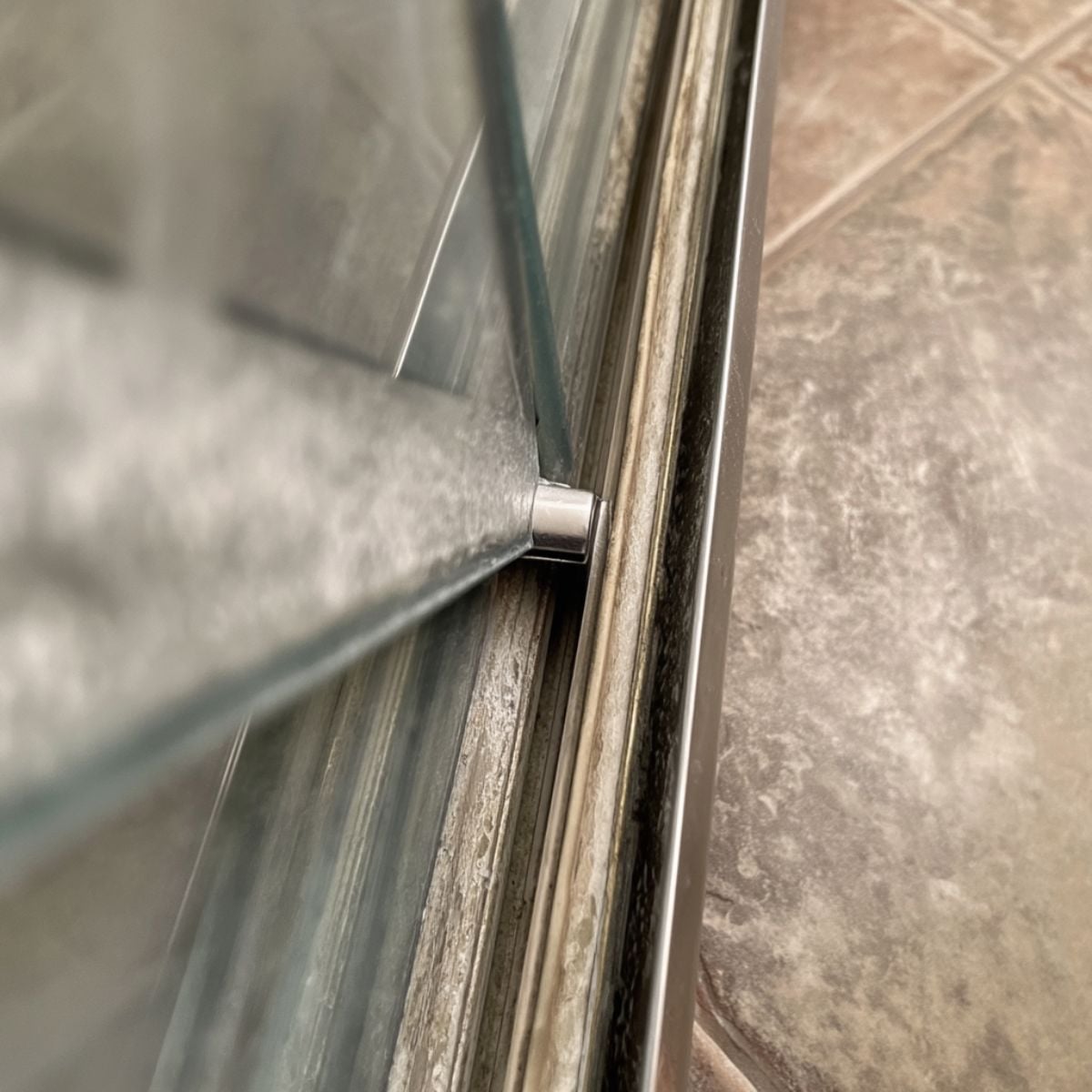 A close up of a glass shower door track showing soap scum buildup, hard water stains, and black mold along the metal frame and floor