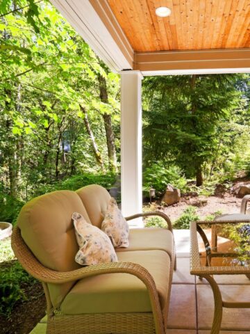 A cozy covered patio with wicker furniture and plush cushions, set against a backdrop of a wooded area. A decorative fountain is visible in the background, adding to the tranquil ambiance.