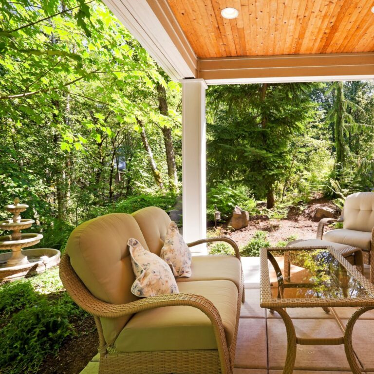 A cozy covered patio with wicker furniture and plush cushions, set against a backdrop of a wooded area. A decorative fountain is visible in the background, adding to the tranquil ambiance.