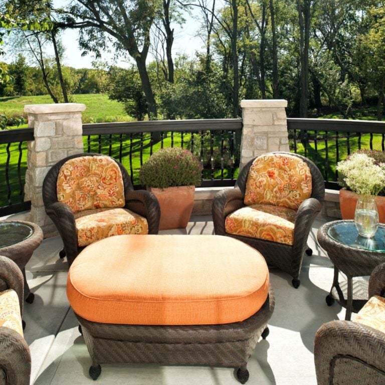 A stylish outdoor seating area with wicker furniture and vibrant floral cushions. The deck features an ottoman and is set against a scenic view of a green landscape, with potted plants adding to the decor.