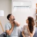 A man and woman sitting on a couch, looking up at an air conditioning unit on the wall, appearing hot and uncomfortable.