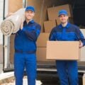 Two movers dressed in blue uniforms are standing in front of an open moving truck filled with cardboard boxes. One mover is holding a rolled-up carpet on his shoulder and smiling, while the other is holding a large cardboard box and looking towards the camera. The scene appears to be outdoors, possibly in a residential area.