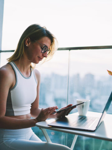 A woman wearing glasses and a workout outfit sits at a small table on a balcony, working on her laptop and using her phone. A potted plant and a cup of coffee are also on the table, with a cityscape visible in the background.