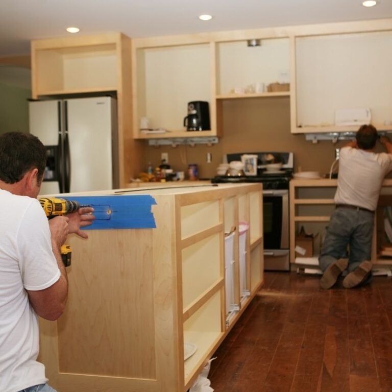 Two men are working on a kitchen remodel. One is drilling into a wooden cabinet with blue tape on it, while the other is installing hardware in the background. The kitchen features an unfinished island, open cabinets, and a refrigerator. The floor is covered with protective material to catch debris.