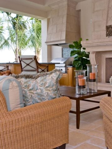 Luxurious outdoor living space featuring wicker furniture with patterned cushions, a stone fireplace, and an outdoor kitchen area with stainless steel appliances and palm trees in the background