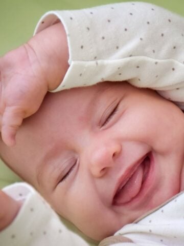 A close-up of a smiling baby lying down, wearing a light-colored onesie with small polka dots, one arm lifted near their head. The baby appears content and joyful with eyes closed and mouth open in a happy expression.