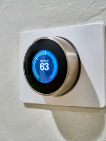 A modern smart thermostat mounted on a white wall, displaying a blue interface with the heat set to 63 degrees Fahrenheit. The sleek, circular design adds a touch of style to the device, making it both functional and visually appealing.