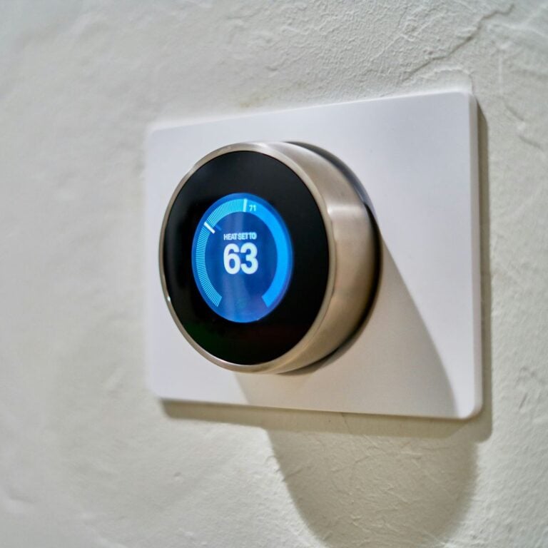 A modern smart thermostat mounted on a white wall, displaying a blue interface with the heat set to 63 degrees Fahrenheit. The sleek, circular design adds a touch of style to the device, making it both functional and visually appealing.