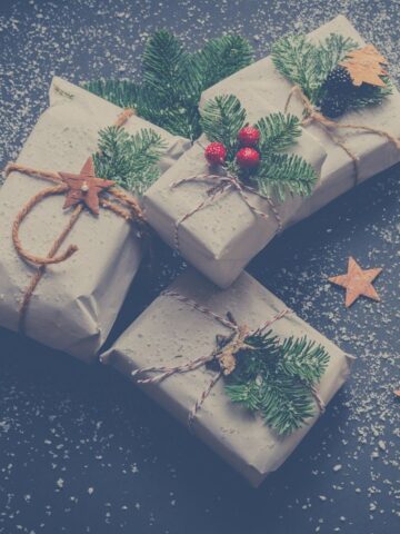 Rustic Christmas presents wrapped in natural-colored paper, decorated with sprigs of evergreen, twine, and small festive embellishments like red berries and wooden stars, scattered on a snowy, dark background with small wooden stars around for a cozy holiday atmosphere.