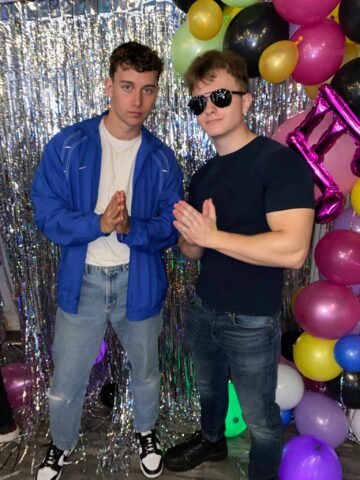 Two young men are posing together in front of a colorful balloon backdrop with metallic streamers. The man on the left is wearing a blue jacket, a white t-shirt, and ripped jeans, while the man on the right is dressed in a dark t-shirt, sunglasses, and dark jeans. They are both making a gesture with their hands, looking confidently at the camera. The background includes a mix of balloons in various colors, creating a lively, festive atmosphere.