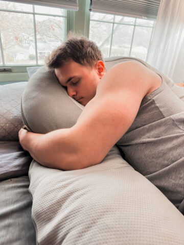 A muscular teenager wearing a tank top is peacefully sleeping on the HUGL Cooling Body Pillow, which wraps around him on a neatly made bed near a window with soft curtains.