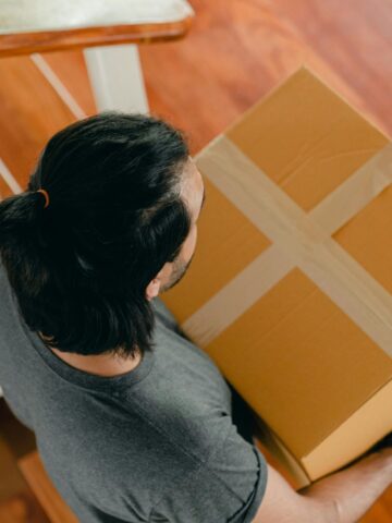 Overhead view of a man with dark hair tied in a ponytail, wearing a gray T-shirt, carrying a taped cardboard shipping box while walking down a wooden staircase.