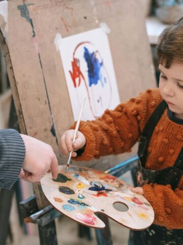 A young child wearing a burnt orange knitted sweater and a paint-stained apron is holding a wooden artist's palette filled with colorful paint. The child is carefully dipping a paintbrush into the palette while standing in front of an easel with a partially completed abstract painting taped to it. An adult’s hand is guiding the child’s brush.