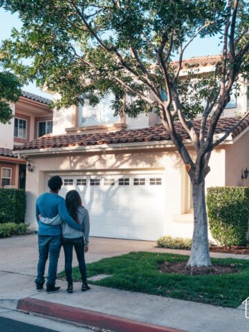 A couple stands arm in arm in front of a two-story suburban house with a garage, admiring the property. The house has a beige stucco exterior, a tiled roof, and lush landscaping with a large tree in the front yard. The couple is seen from behind, standing on the sidewalk.
