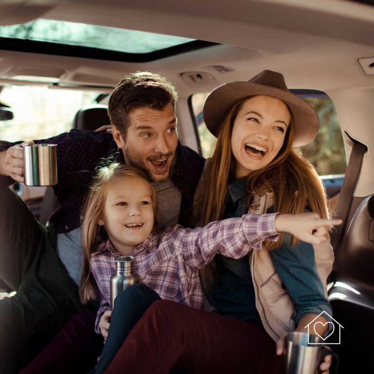 A cheerful family of three sits inside an SUV during a road trip, holding thermoses and laughing together. The young daughter excitedly points outside, while the fall scenery is visible through the car windows.