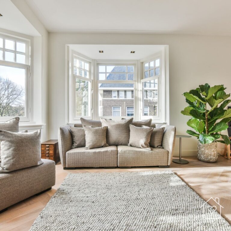 A bright and airy living room with large bay windows, a neutral-toned sofa adorned with matching throw pillows, a textured area rug, and a lush fiddle-leaf fig plant in a decorative planter. The space features a modern, minimalist aesthetic with natural light streaming in through the windows.