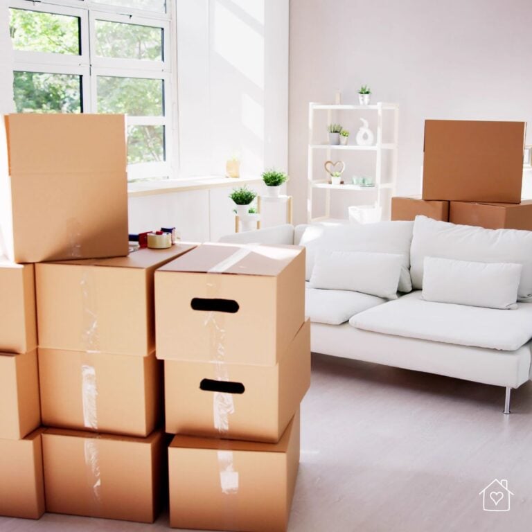 A well-lit living room with neatly stacked cardboard moving boxes, packing tape, and a white couch, ready for a move. A minimalist shelving unit with small plants adds a cozy touch to the space.