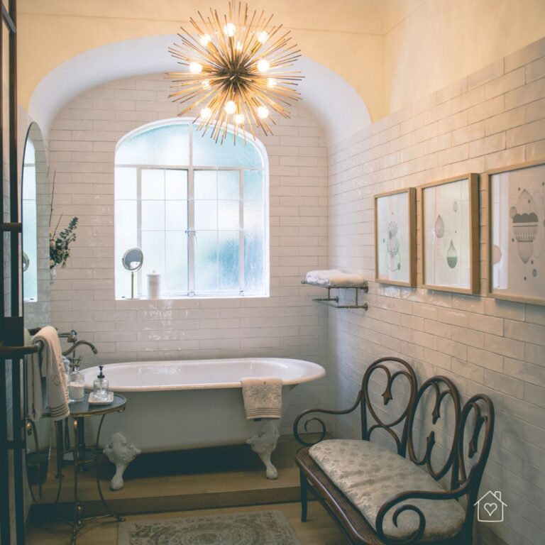 A beautifully designed bathroom featuring a classic clawfoot bathtub, white subway tile walls, and an arched window. A striking gold starburst pendant light hangs from the ceiling, adding a touch of elegance and warmth.