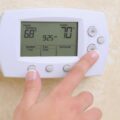 A hand pressing a button on a digital thermostat, demonstrating how setting the right indoor temperature can keep a home comfortable in all weather conditions.