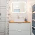 A bright and airy small bathroom featuring a backlit LED mirror, a modern white sink, and a wooden countertop. The tiled walls and soft lighting make the space feel more open.