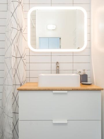 A bright and airy small bathroom featuring a backlit LED mirror, a modern white sink, and a wooden countertop. The tiled walls and soft lighting make the space feel more open.