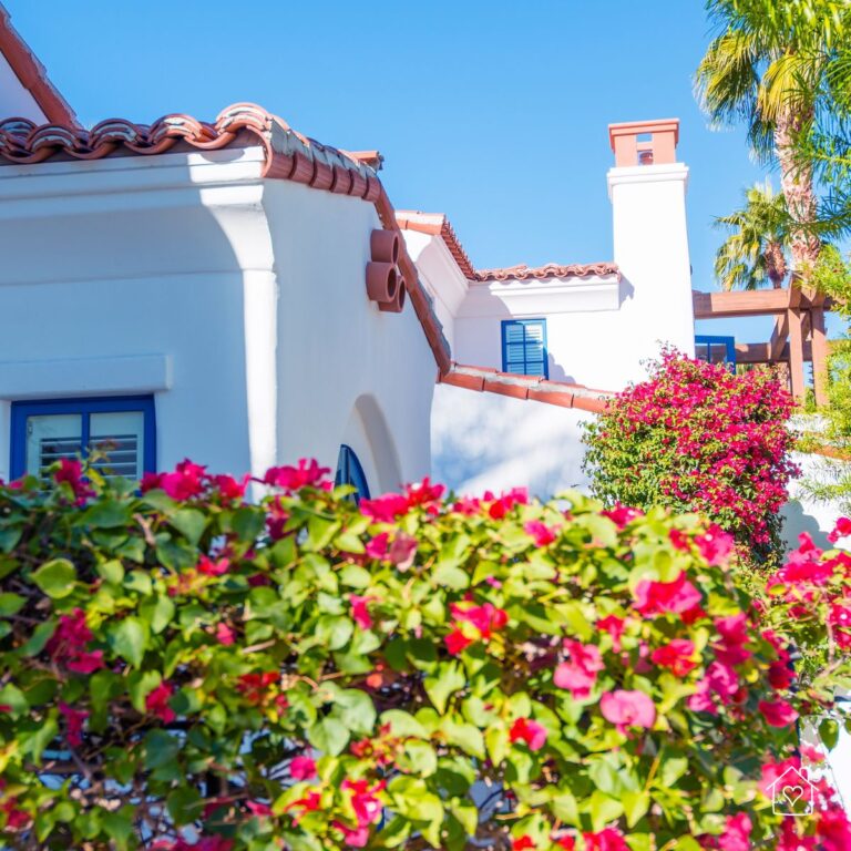 A stunning Mediterranean-style home with white stucco walls, terracotta roof tiles, and bright pink bougainvillea in full bloom. The vibrant colors and lush greenery create a quintessential Mediterranean ambiance.