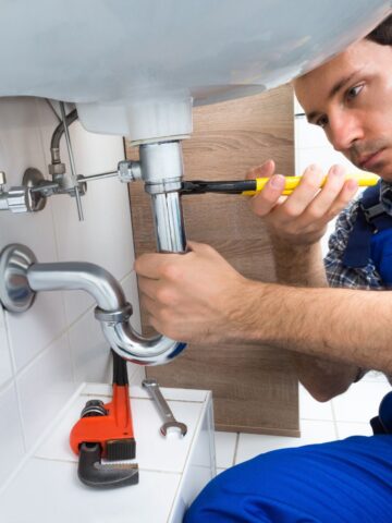 Professional plumber in blue overalls using tools to fix a bathroom sink drain, with plumbing wrenches on the counter.