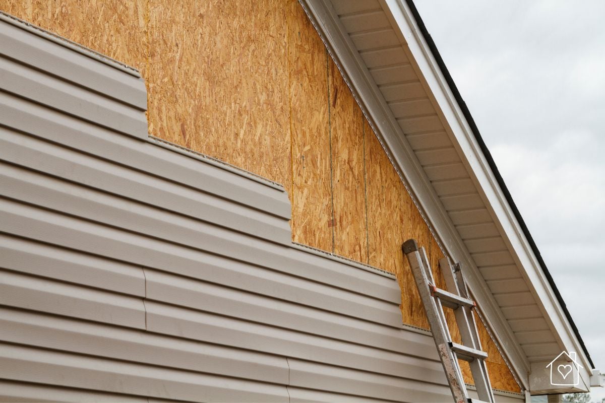 A work-in-progress vinyl siding installation, showing a partially completed wall with exposed plywood sheathing and a ladder leaning against the house, demonstrating the siding application process.