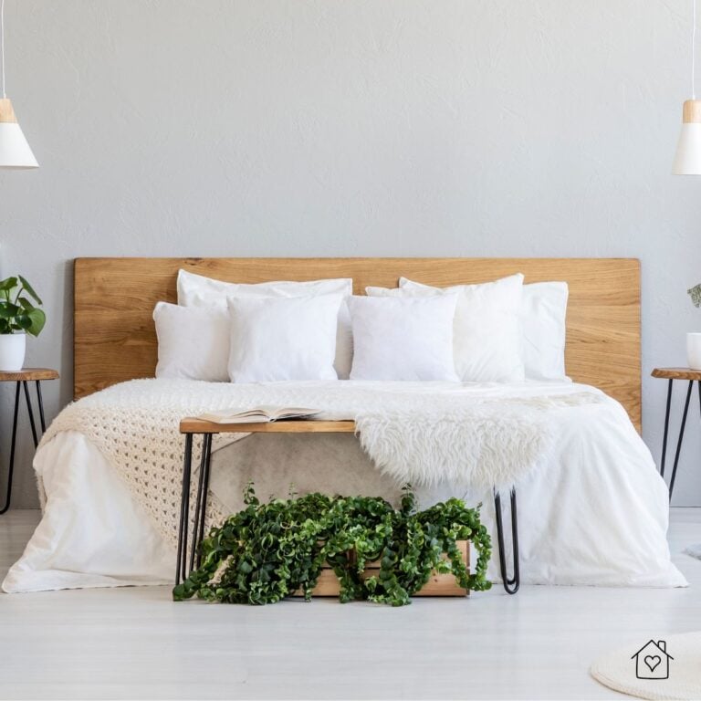 A cozy bedroom with a wooden headboard, crisp white bedding, and a plush faux fur blanket. A small wooden bench with a lush green plant sits at the foot of the bed, adding a touch of nature to the neutral space.