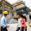 A general contractor wearing a yellow hard hat explains construction details to a couple in front of a modern home under renovation.