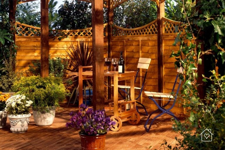 Intimate wooden deck setup with a small table, chairs, and wine glasses surrounded by planters and a lattice privacy fence for a warm, enclosed garden feel.