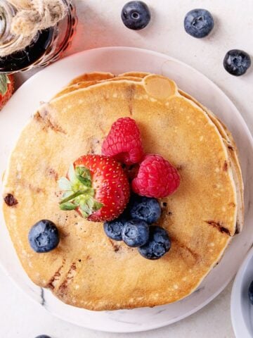 Overhead view of warm pancakes topped with fresh strawberries, raspberries, and blueberries, surrounded by more fruit and syrup—an inviting setup for a family pancake morning.