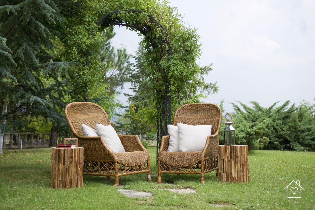 Two wicker chairs with white pillows sit beneath a leafy arch in a peaceful backyard seating area.