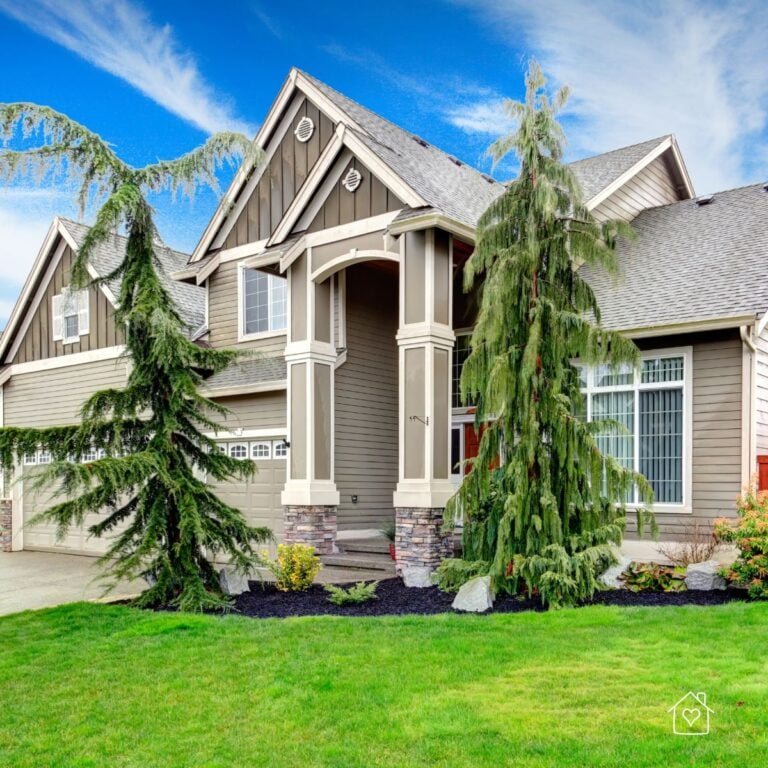 Striking Craftsman-style house with taupe vertical siding, tall pillars, and steep rooflines framed by evergreen trees.