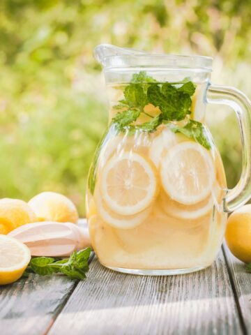 Glass pitcher filled with refreshing lemon water and mint leaves, sitting on a rustic wooden table outdoors, surrounded by fresh lemons — a perfect way to stay hydrated and energized throughout a busy day.