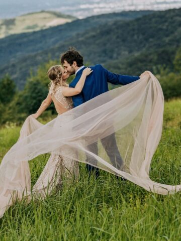 Newlyweds embracing in a lush green meadow with mountains in the background, representing a romantic outdoor wedding setting.