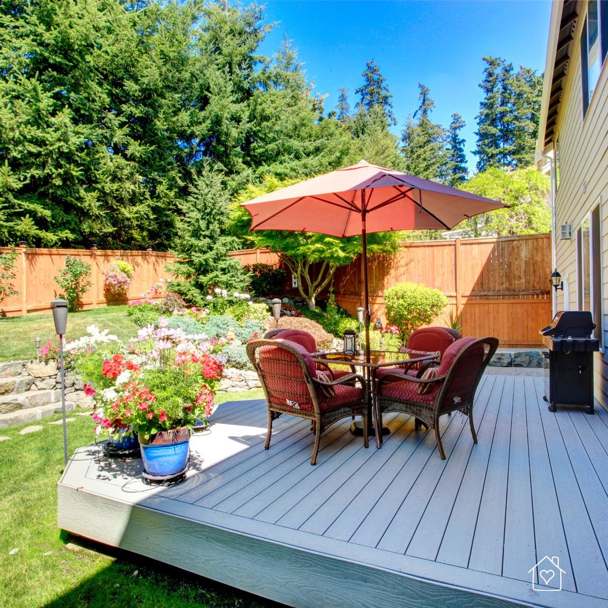 Raised backyard deck featuring a round dining table with four wicker chairs, a red umbrella, and colorful potted flowers, surrounded by lush landscaping and a wooden privacy fence.