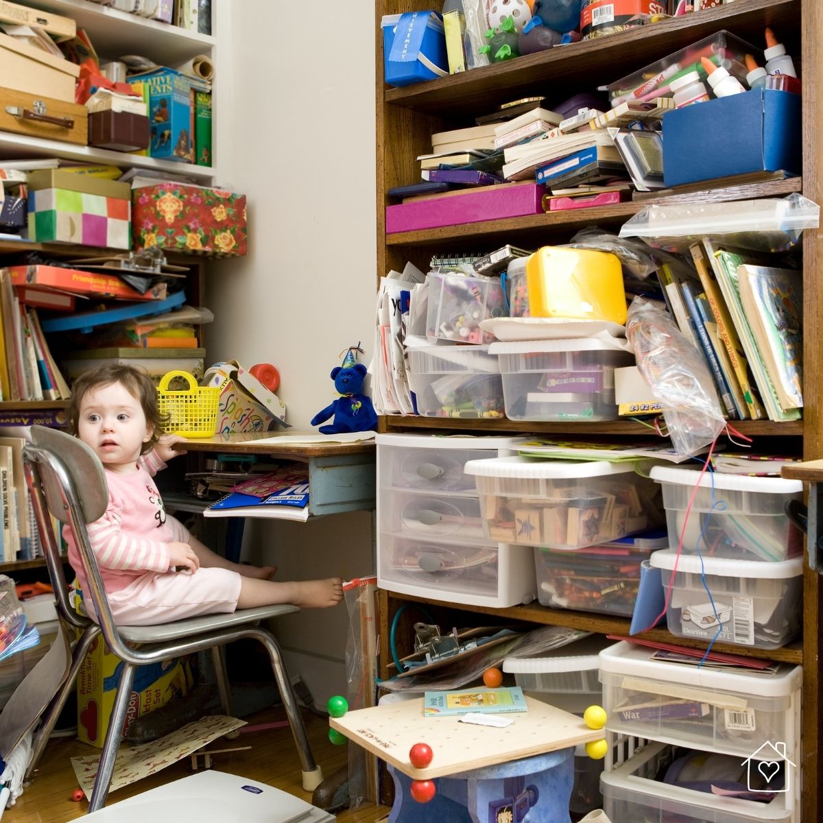 A young child sits in a cluttered room surrounded by overflowing shelves, disorganized bins, and piles of papers and toys—highlighting the chaos before a home makeover.