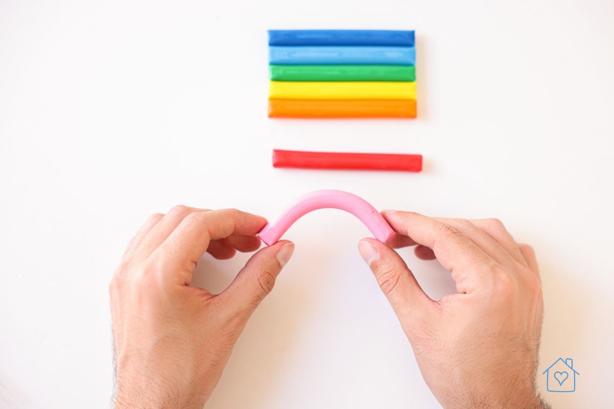 Hands bending a pink piece of polymer clay with a row of bright clay sticks in the background, demonstrating the pliability of beginner-friendly clay.