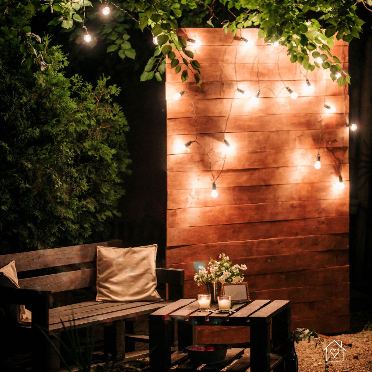 Cozy nighttime patio setup with a wooden bench, pallet-style coffee table, glowing string lights on a wood backdrop, and decorative candles and flowers for a warm, inviting ambiance.