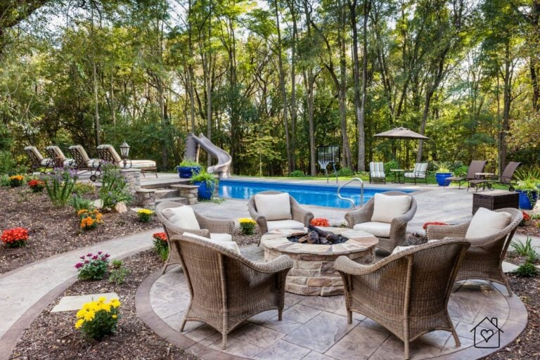 Outdoor living area featuring a stamped tile patio around a stone fire pit and pool, surrounded by lush landscaping and wicker seating.