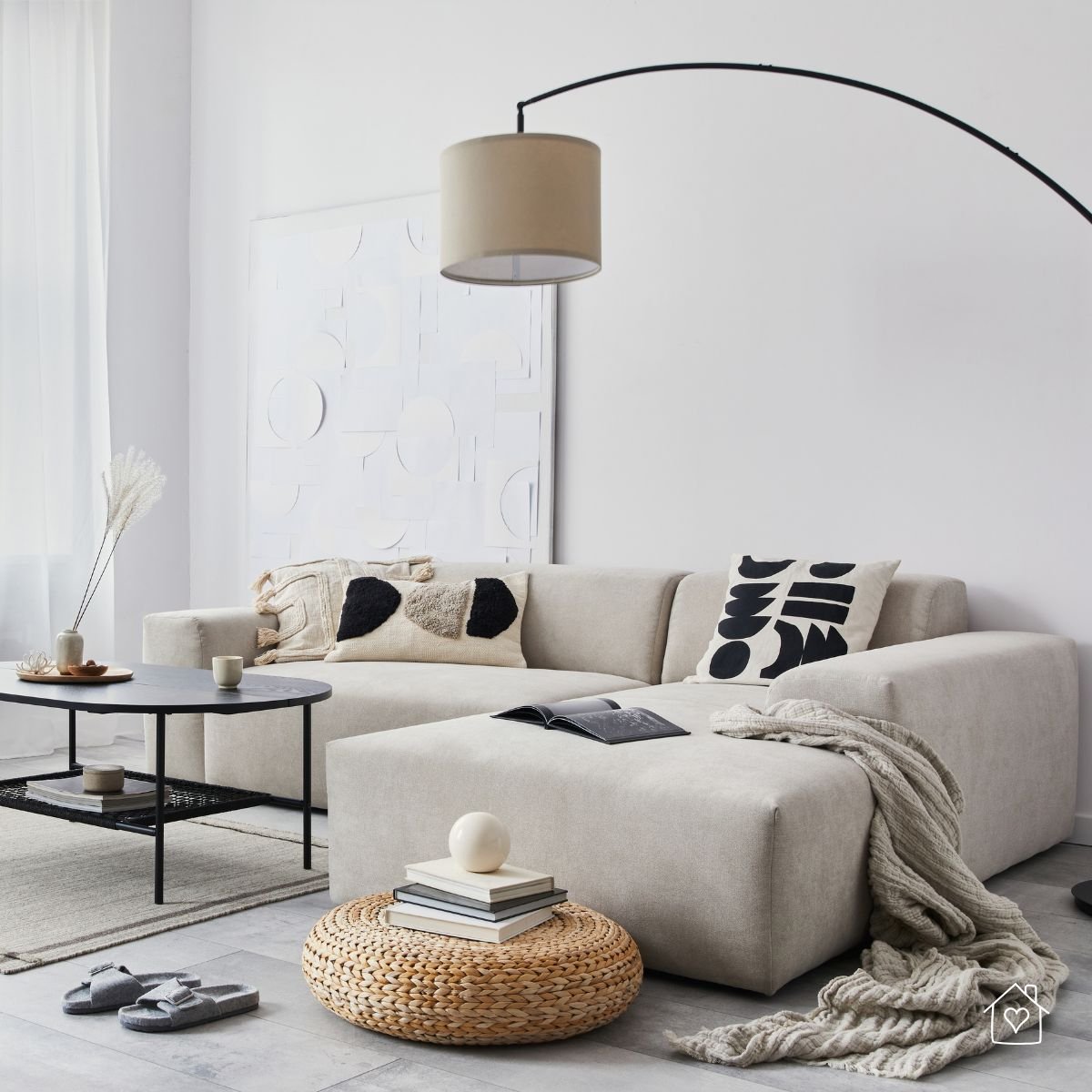 A neutral-toned living room with a plush sectional sofa, abstract pillows, a round wicker pouf, and cozy throws creating a modern yet inviting space.
