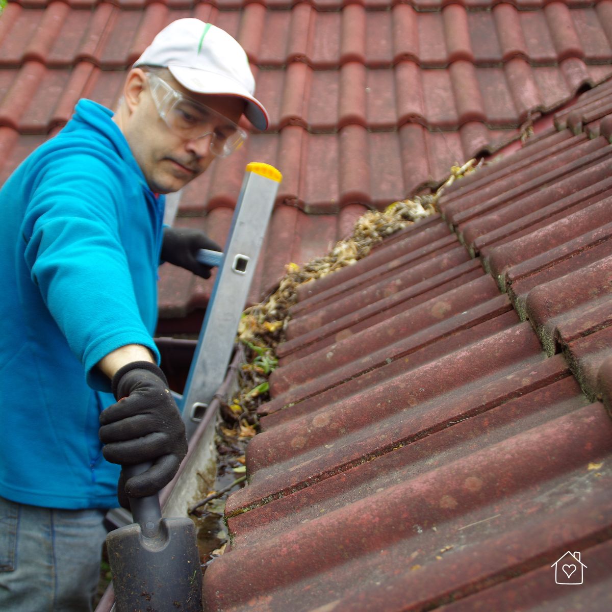 A man wearing protective gloves and safety goggles removes debris from a clogged rain gutter on a tiled roof using a scoop.