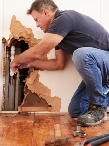 Man kneeling on hardwood floor fixing pipes inside a severely damaged wall during an emergency home repair.