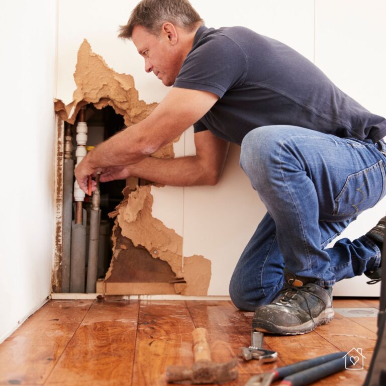 Man kneeling on hardwood floor fixing pipes inside a severely damaged wall during an emergency home repair.
